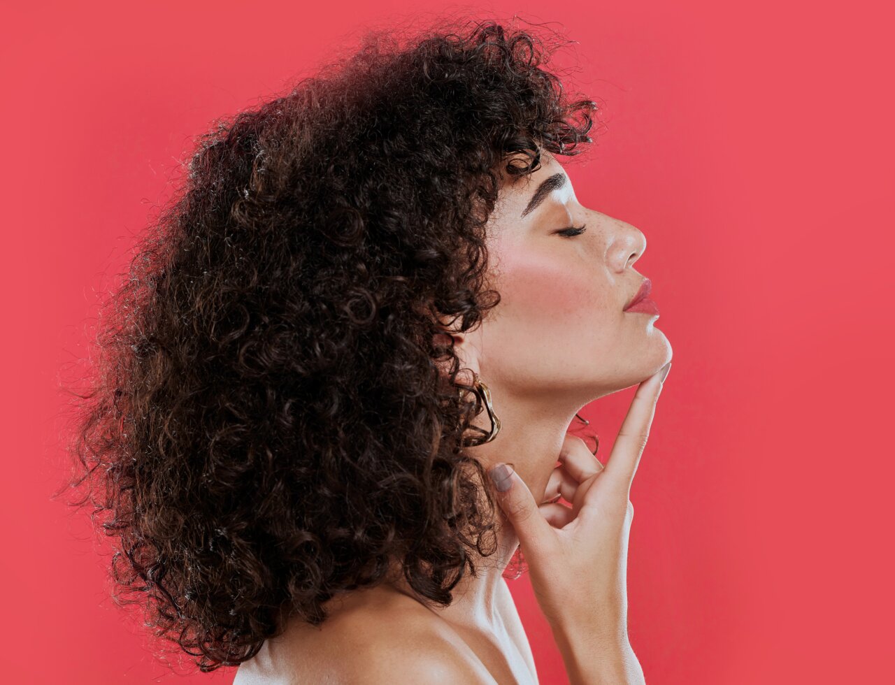 Junge schöne Frau mit lockigem Haar und rosigen Wangen vor einem pinken Hintergrund als Sinnbild für das Trendmakeup 2025 Workout Blush